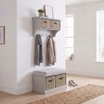 A truffle-coloured wall-mounted shelf unit displaying a framed print and potted plant on top, with two natural woven baskets featuring dark handles in its cubbies and four silver double-hooks holding a grey scarf and a beige coat. Directly below sits a matching truffle wooden storage bench topped with a light grey cushion, containing two larger natural woven baskets in its base. The furniture is styled in a bright hallway with light wood flooring, a woven runner rug, and a pair of canvas sneakers resting beside the bench.
