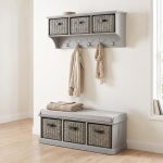 A light grey wall-mounted shelf unit with three grey woven baskets featuring dark handles, and five silver double-hooks holding a light beige jacket and a fringed scarf. Directly below sits a matching grey wooden storage bench topped with a light grey cushion and a folded beige knitted blanket, containing three larger matching grey woven baskets in its base. The furniture is styled in a bright hallway with light wood flooring against an off-white wall