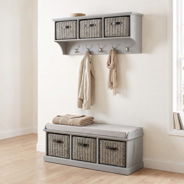 A light grey wall-mounted shelf unit with three grey woven baskets featuring dark handles, and five silver double-hooks holding a light beige jacket and a fringed scarf. Directly below sits a matching grey wooden storage bench topped with a light grey cushion and a folded beige knitted blanket, containing three larger matching grey woven baskets in its base. The furniture is styled in a bright hallway with light wood flooring against an off-white wall