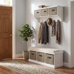 A white wall-mounted shelf unit displaying small decorative boxes on top, with three natural woven baskets featuring dark handles. Five silver double-hooks below hold a tan jacket, a dark jacket, a straw hat, and two patterned scarves. Directly below sits a matching white wooden storage bench topped with a beige cushion, containing three larger matching natural woven baskets in its base. The furniture is styled in a bright entryway next to a wooden door and a potted fiddle leaf fig plant, with wood flooring and a patterned runner rug.