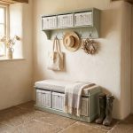 A sage green wall-mounted shelf unit with three white woven baskets featuring small white bow details in its open compartments. Five silver double-hooks below hold a canvas tote bag, a straw hat with a black band, and a dried floral wreath. Directly below sits a matching sage green wooden storage bench topped with a beige cushion and a draped patterned fringed blanket, containing three larger matching white woven baskets in its base. The furniture is styled in a rustic room with uneven plaster walls, a stone flagstone floor, and a pair of tall green Wellington boots resting beside the bench.
