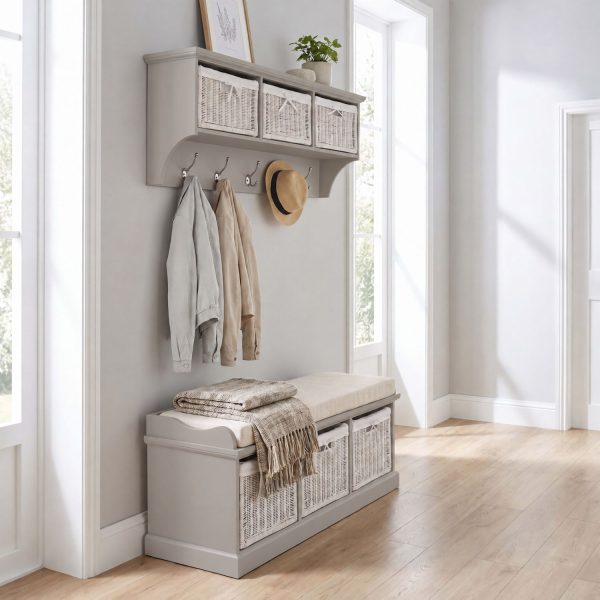 A truffle-coloured wall-mounted shelf unit displaying a framed print and potted plant on top, with three white woven baskets featuring small white bow details in its open compartments. Five silver double-hooks below hold a light grey jacket, a tan jacket, and a straw hat. Directly below sits a matching wooden storage bench topped with a beige cushion and a folded patterned fringed blanket, containing three larger matching white woven baskets in its base. The furniture is styled in a bright hallway with light wood flooring and soft grey walls.