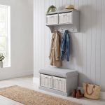 A grey wall-mounted shelf unit displaying a potted trailing plant and a straw hat on top, with two white woven baskets featuring small white bow details in its open compartments. Four silver double-hooks below hold a beige trench coat and a blue denim jacket. Directly below sits a matching grey wooden storage bench topped with a light grey cushion, containing two larger matching white woven baskets in its base. The furniture is styled in a bright hallway against a white panelled wall, with light wood flooring, a woven runner rug, and a pair of brown leather boots alongside a straw tote bag resting beside the bench.