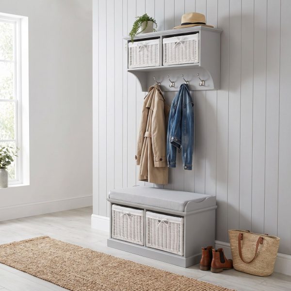 A grey wall-mounted shelf unit displaying a potted trailing plant and a straw hat on top, with two white woven baskets featuring small white bow details in its open compartments. Four silver double-hooks below hold a beige trench coat and a blue denim jacket. Directly below sits a matching grey wooden storage bench topped with a light grey cushion, containing two larger matching white woven baskets in its base. The furniture is styled in a bright hallway against a white panelled wall, with light wood flooring, a woven runner rug, and a pair of brown leather boots alongside a straw tote bag resting beside the bench.