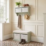 A sage green wall-mounted shelf unit displaying a potted fern, leather gloves, and a small brown box on top, with two white woven baskets featuring small white bow details in its open compartments. Four silver double-hooks below hold a draped beige scarf and a straw hat with a black band. Directly below sits a matching sage green wooden storage bench topped with a beige cushion, containing two larger matching white woven baskets in its base. The furniture is styled in a bright entryway with cream panelled walls, patterned geometric floor tiles, and a pair of brown leather lace-up boots resting in front of the bench