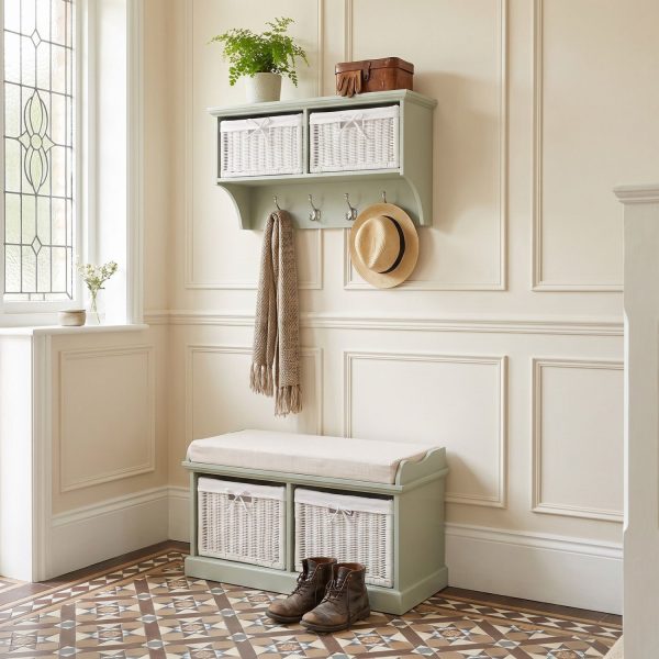 A sage green wall-mounted shelf unit displaying a potted fern, leather gloves, and a small brown box on top, with two white woven baskets featuring small white bow details in its open compartments. Four silver double-hooks below hold a draped beige scarf and a straw hat with a black band. Directly below sits a matching sage green wooden storage bench topped with a beige cushion, containing two larger matching white woven baskets in its base. The furniture is styled in a bright entryway with cream panelled walls, patterned geometric floor tiles, and a pair of brown leather lace-up boots resting in front of the bench