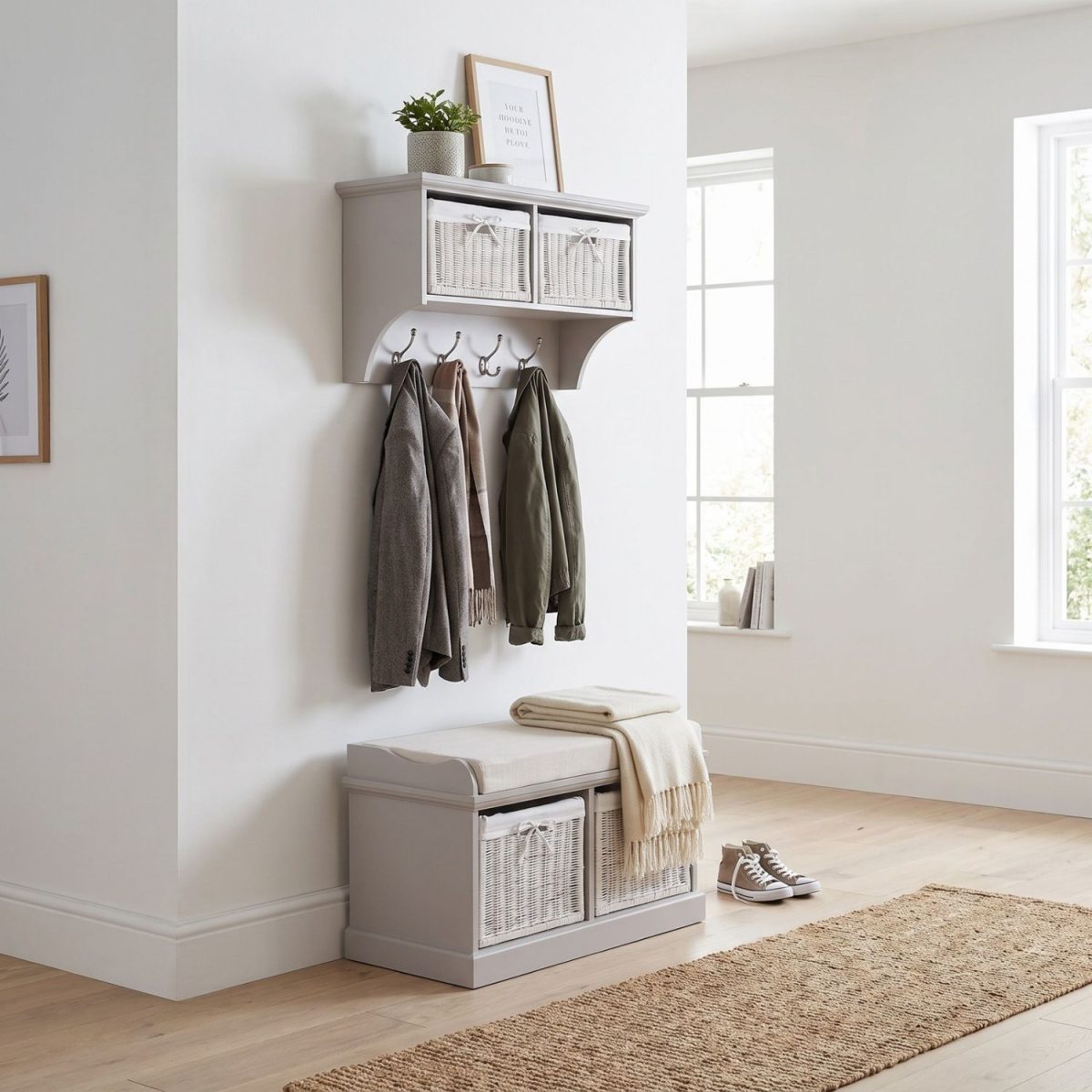 A truffle-coloured wall-mounted shelf unit displaying a framed print and potted plant on top, with two white woven baskets featuring small white bow details in its open compartments. Four silver double-hooks below hold a grey jacket, a brown scarf, and an olive green jacket. Directly below sits a matching wooden storage bench topped with a beige cushion and a folded cream fringed blanket, containing two larger matching white woven baskets in its base. The furniture is styled in a bright hallway with light wood flooring, a woven runner rug, and a pair of canvas sneakers resting beside the bench.
