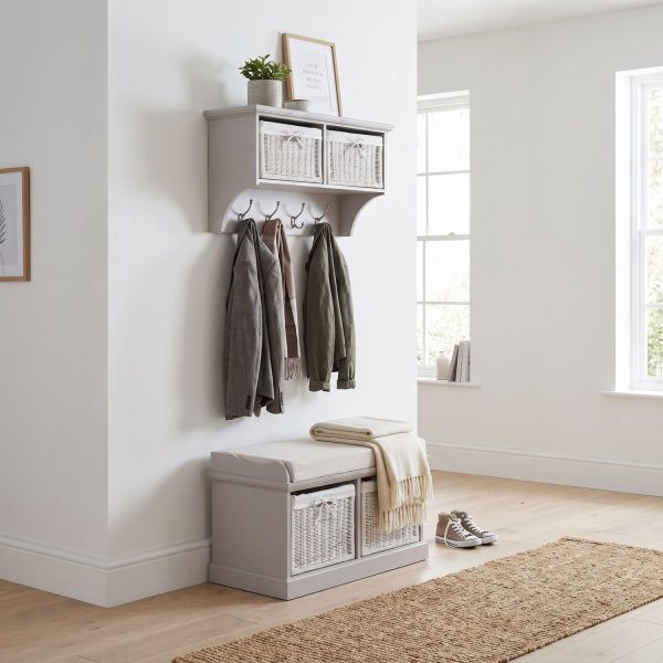 A truffle-coloured wall-mounted shelf unit displaying a framed print and potted plant on top, with two white woven baskets featuring small white bow details in its open compartments. Four silver double-hooks below hold a grey jacket, a brown scarf, and an olive green jacket. Directly below sits a matching wooden storage bench topped with a beige cushion and a folded cream fringed blanket, containing two larger matching white woven baskets in its base. The furniture is styled in a bright hallway with light wood flooring, a woven runner rug, and a pair of canvas sneakers resting beside the bench.