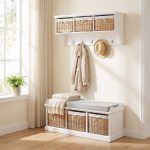 A white wall-mounted shelf unit with three natural woven baskets featuring white cloth liners and bow details in its open compartments. Five silver double-hooks below hold a beige trench coat and a straw hat. Directly below sits a matching white wooden storage bench topped with a grey cushion and a folded cream fringed blanket, containing three larger matching natural woven baskets in its base. The furniture is styled in a bright room next to a window with sheer curtains and a potted fern on the sill, set against cream walls and light wood flooring.