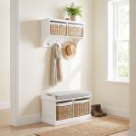 A white wall-mounted shelf unit displaying a potted fern and a small round box on top, with two natural woven baskets featuring white cloth liners and bow details in its open compartments. Four silver double-hooks below hold a draped beige scarf and a straw hat. Directly below sits a matching white wooden storage bench topped with a grey cushion, containing two larger matching natural woven baskets in its base. The furniture is styled in a bright entryway next to a window, set against cream walls with light wood flooring, a woven runner rug, and a pair of brown leather lace-up boots resting beside the bench