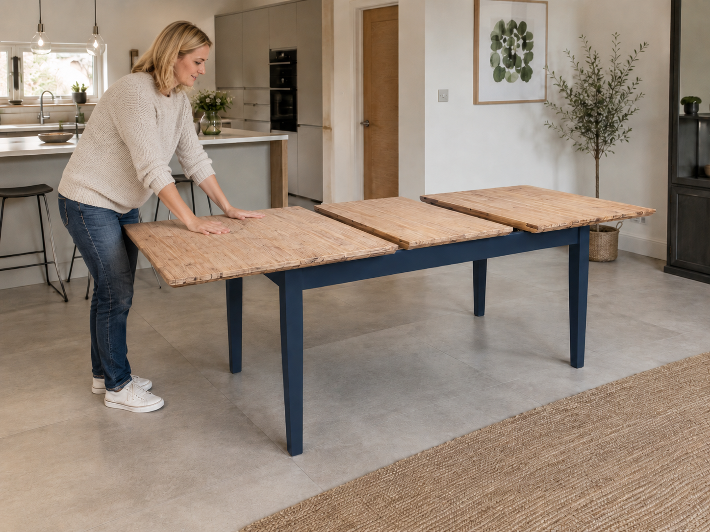 A woman in a modern UK kitchen pushes the wooden tabletop panels of a navy blue extendable dining table together after inserting the centre extension leaf.