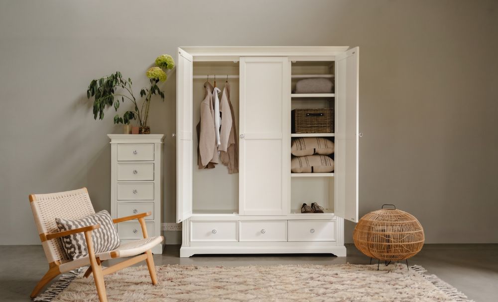 A large white wooden Gainsborough triple wardrobe with three drawers at the base, shown in a bedroom setting with other white furniture including a tallboy and an armchair, all on a patterned rug.