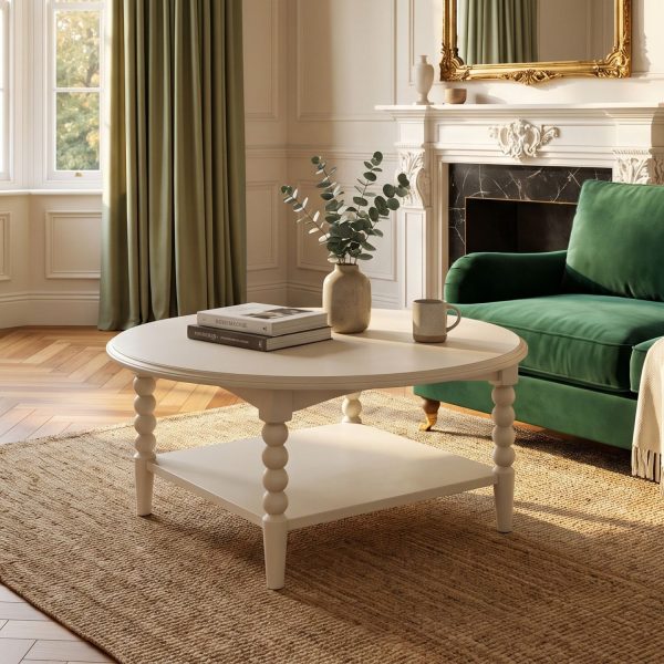 A linen-coloured round coffee table characterized by its distinctive bobbin-turned legs and a lower square shelf. The tabletop is styled with two stacked books, a textured vase holding eucalyptus stems, and a ceramic mug. The table is positioned on a large woven rug over a wood herringbone floor, situated in a sunlit living room in front of a dark green velvet sofa and an ornate white fireplace.