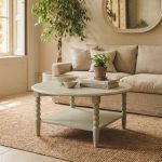 A pale olive round coffee table characterized by its distinctive bobbin-turned legs and a lower square shelf. The tabletop is styled with two stacked books supporting a small potted fern, alongside two light-coloured nesting bowls. The table is positioned on a large woven rug over a light stone floor, situated in a sunlit living room in front of a beige sofa and next to a large potted indoor tree with a braided trunk.