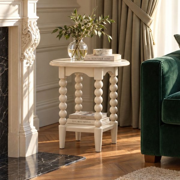 A linen-coloured round side table characterized by its distinctive bobbin-turned legs, arched apron, and a lower square shelf. The tabletop is styled with a ribbed glass vase holding leafy branches, a book titled "The Kinfolk Home", and a small round marble dish. The lower shelf holds two stacked books titled "At Home" and "Live Beautifully". The table is positioned in a sunlit room on a wood herringbone floor, situated between an ornate white fireplace with a black marble hearth and a dark green velvet sofa.