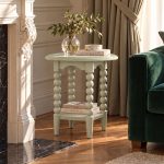 A pale olive round side table characterized by its distinctive bobbin-turned legs, arched apron, and a lower square shelf. The tabletop is styled with a ribbed glass vase holding leafy branches, a book titled "The Kinfolk Home", and a small round marble dish. The lower shelf holds two stacked books titled "At Home" and "Live Beautifully". The table is positioned in a sunlit room on a wood herringbone floor, situated between an ornate white fireplace with a black marble hearth and a dark green velvet sofa.